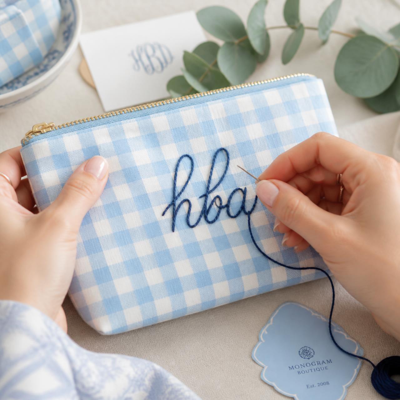 Hands embroidering a navy script monogram onto a powder-blue gingham pouch